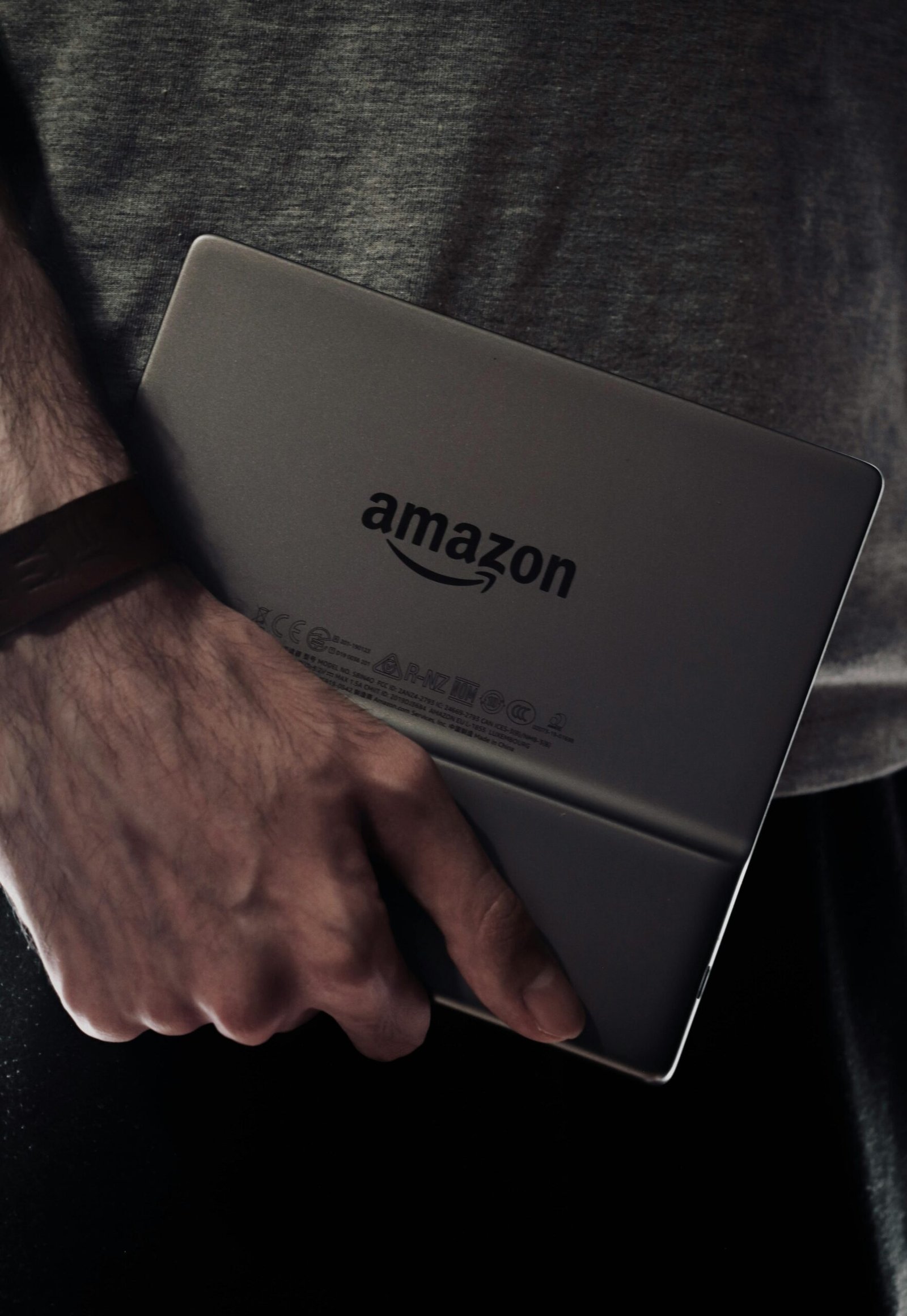 Close-up of a man's hand holding an Amazon Kindle device in a dimly lit setting.