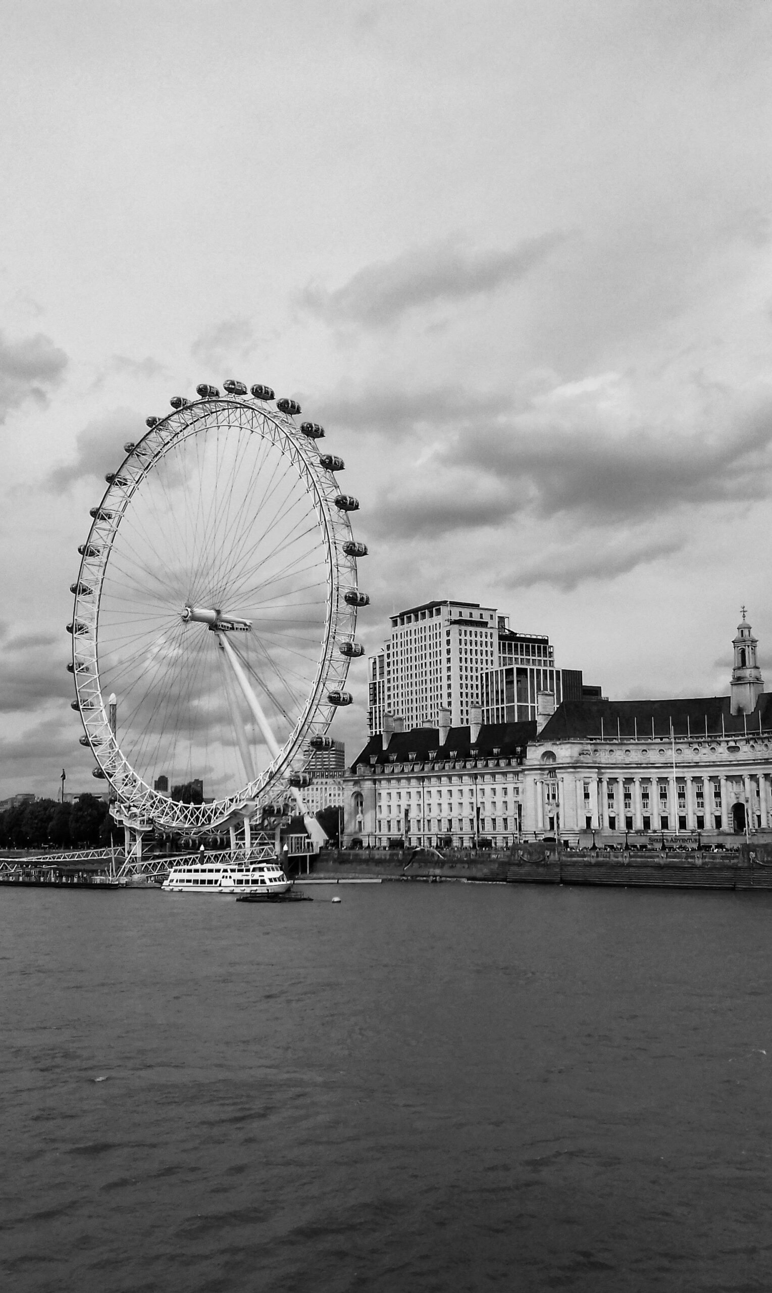 Black and white photo of the iconic London Eye ferris wheel by the River Thames with historic architecture in the background.