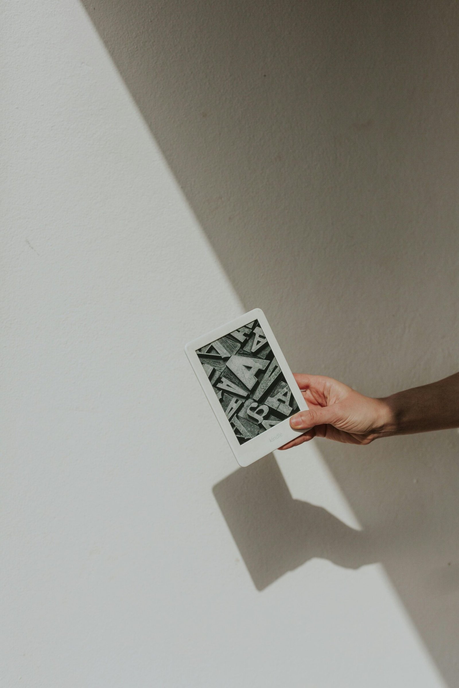 A minimalist photo featuring a hand holding a tablet against a light background, casting a shadow.