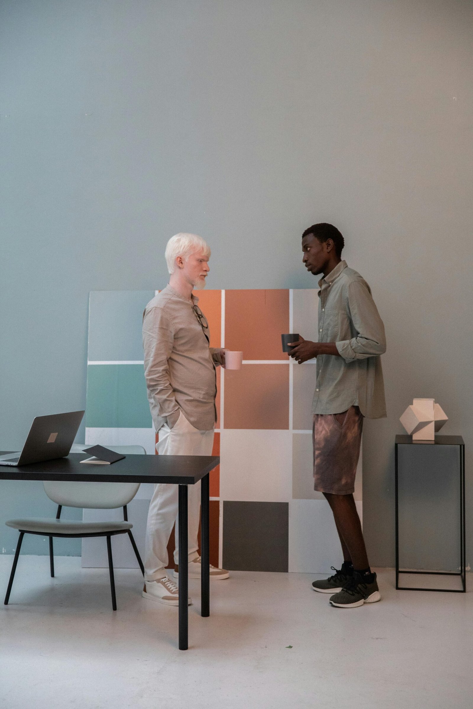 Side view of multiracial coworkers in casual clothes with mugs standing in modern office near picture with color palette and discussing results